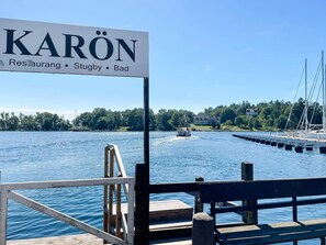 A signboard with 'KARÖN' and amenities listed, a wooden dock, a boat on water, and a distant house.
