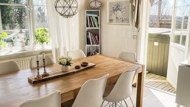 A dining area with a wooden table, white chairs, a bookshelf, and a window with curtains. - 4 Star Holiday Home in Ronneby (Ronneby)