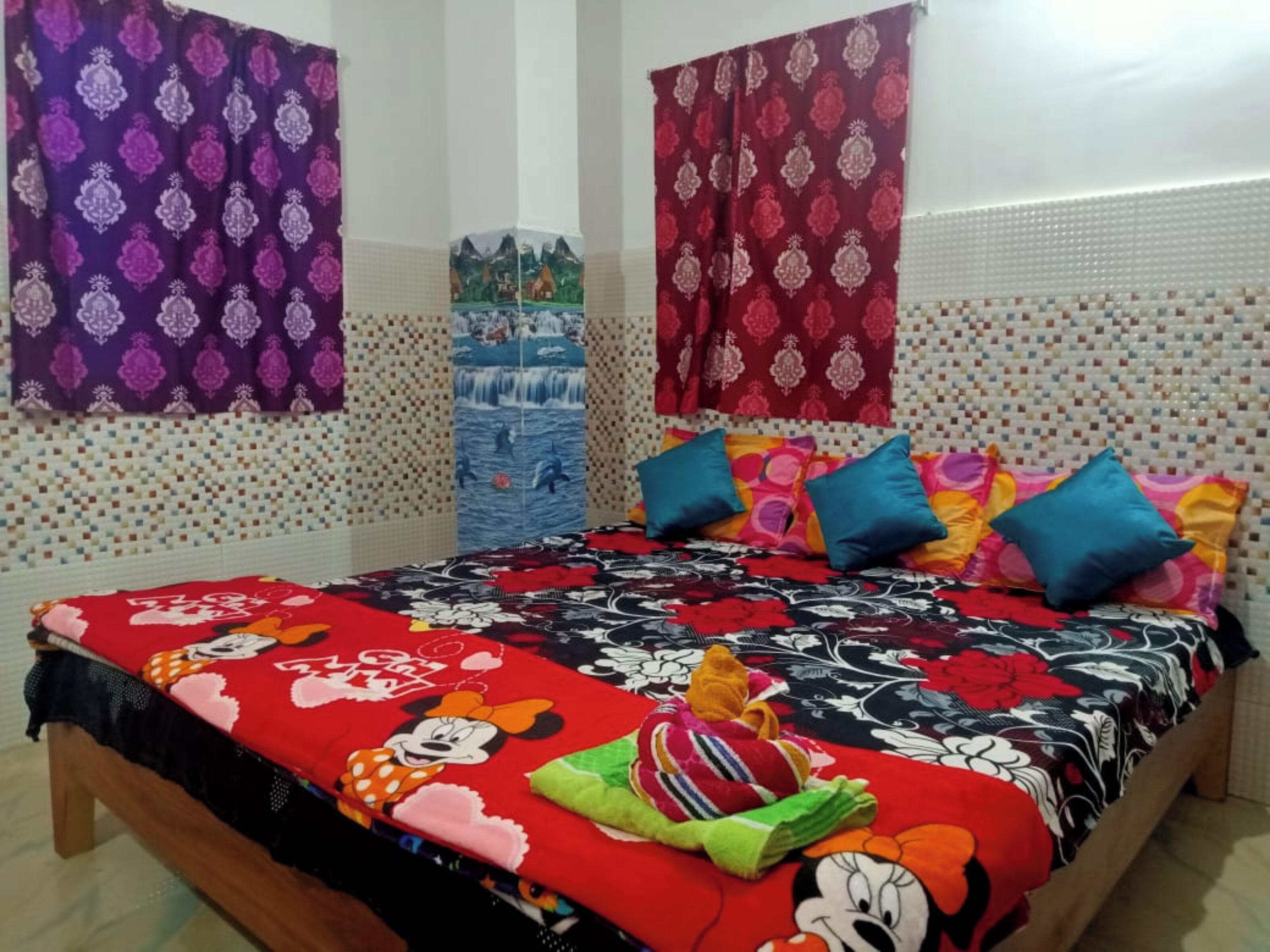 A bedroom with a bed covered in a colorful floral bedspread, a Mickey Mouse pillow, and a Mickey Mouse plush toy. There are two curtains with patterned designs, a wall with a mosaic tile pattern, and a small decorative item on the wall.