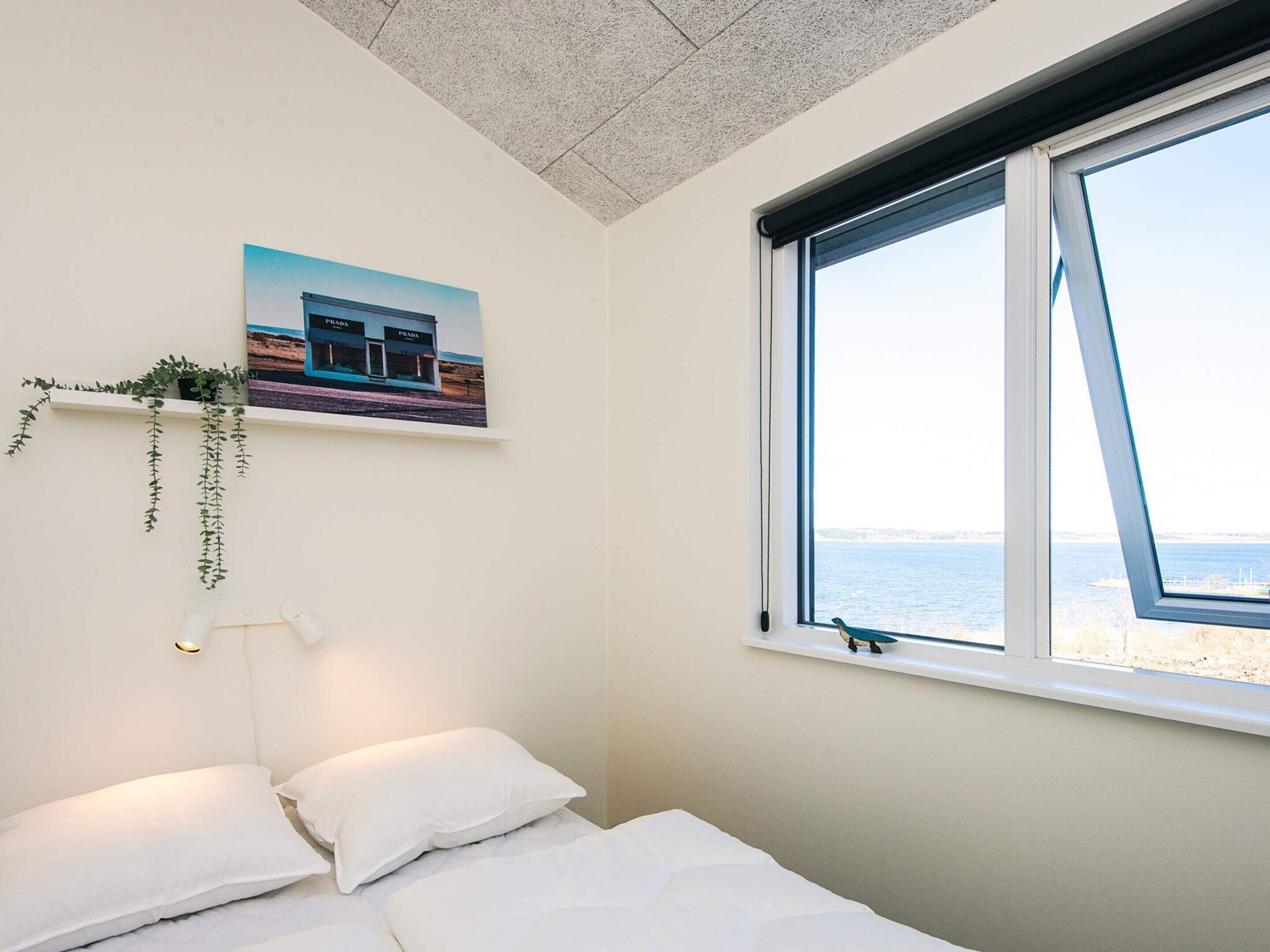 A bedroom with a bed, a window with a view of the sea, a wall-mounted shelf with a plant, and a framed picture of a building.