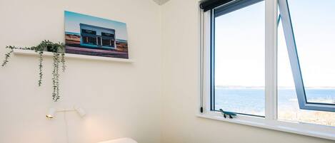 A bedroom with a bed, a window with a view of the sea, a wall-mounted shelf with a plant, and a framed picture of a building.