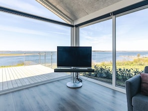 A modern living room with a large TV on a stand, a grey sofa, and a view of a lake through large windows. - 5 Star Holiday Home in Logstrup (Logstrup)