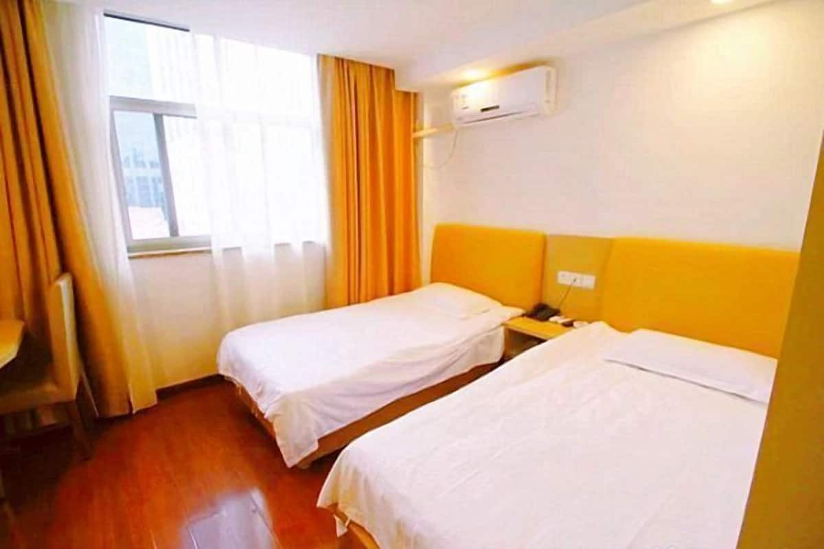 A hotel room with two single beds, a desk, a chair, a window with curtains, and an air conditioning unit.