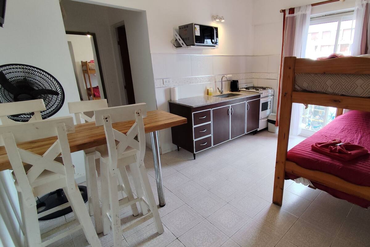 A compact room with a bunk bed, a small kitchenette, a dining table, and a fan.