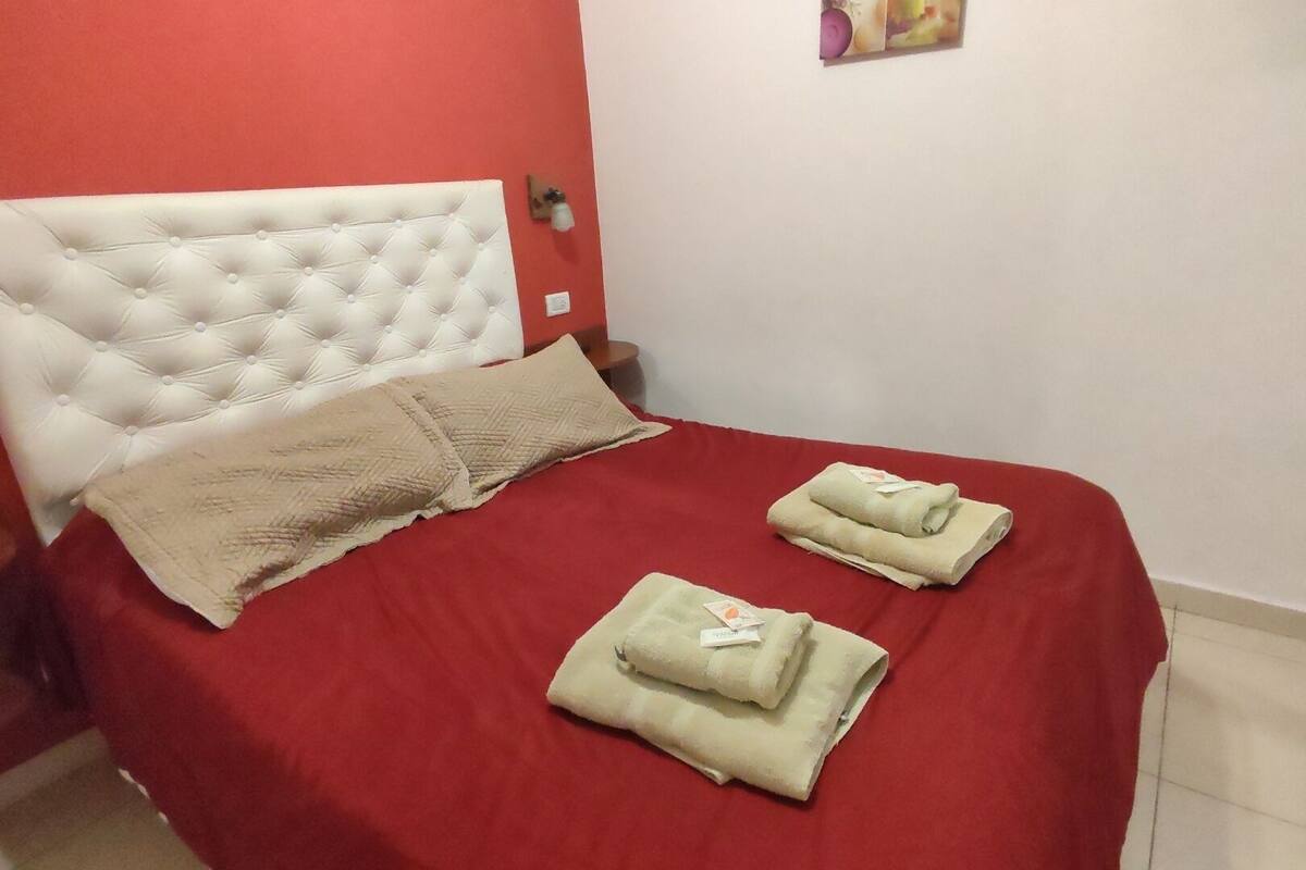 A neatly made bed with a red bedspread and two folded towels, a white tufted headboard, and a small bedside table with a lamp.