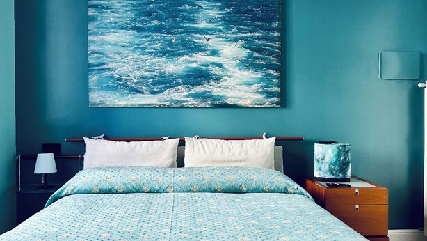 Une chambre à coucher avec un lit, des oreillers, une table de chevet, une lampe et un tableau représentant l’océan.