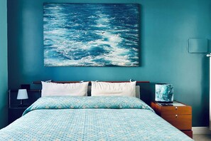 Ein Schlafzimmer mit Bett, Kissen, einem Nachttisch, einer Lampe und einem Meeresbild.