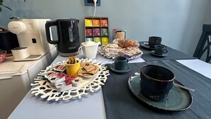 Une table dressée pour le petit-déjeuner, avec une machine à café, des sachets de thé, des viennoiseries et des tasses.