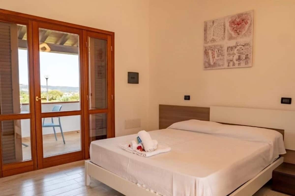 A hotel room with a bed, a small table with a towel and toiletries, a balcony with a chair, and a wall with a decorative piece.