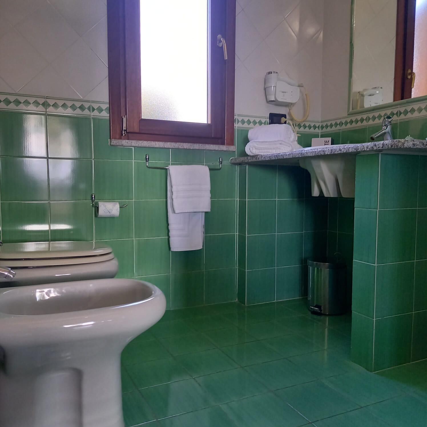 Et badeværelse med grønne fliser, et toilet, en håndvask og et vindue.