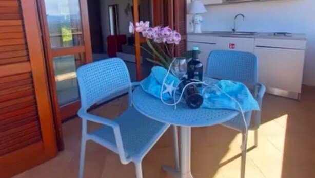 A room with a sliding glass door leading to a balcony, a small round table with chairs, a vase of flowers, and a kitchenette with a sink and cabinets. - My Tiffany Rooms (Tanaunella)