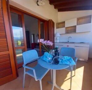 A room with a sliding glass door leading to a balcony, a small round table with chairs, a vase of flowers, and a kitchenette with a sink and cabinets. - My Tiffany Rooms (Tanaunella)