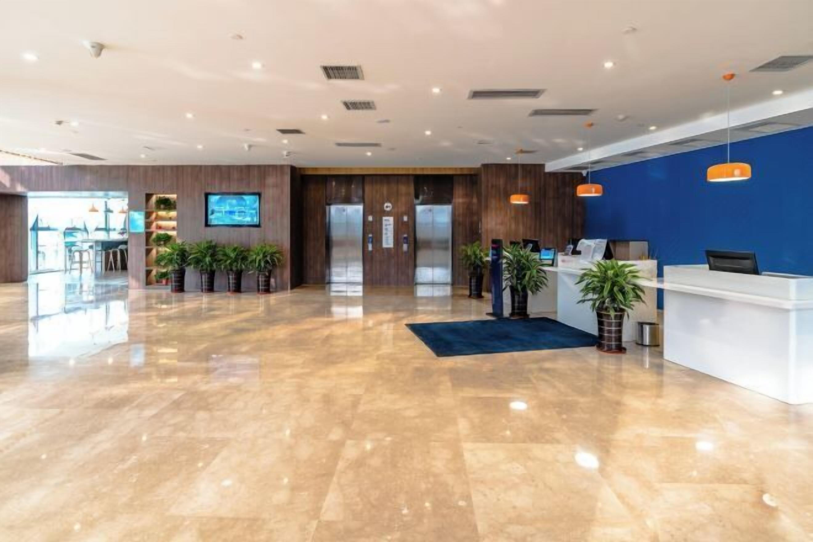 A modern hotel lobby with a reception desk, elevators, and a large screen.