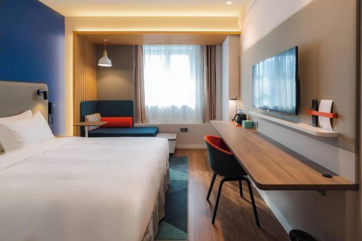 Una habitación de hotel con cama, escritorio, silla, televisor y ventana con cortinas.