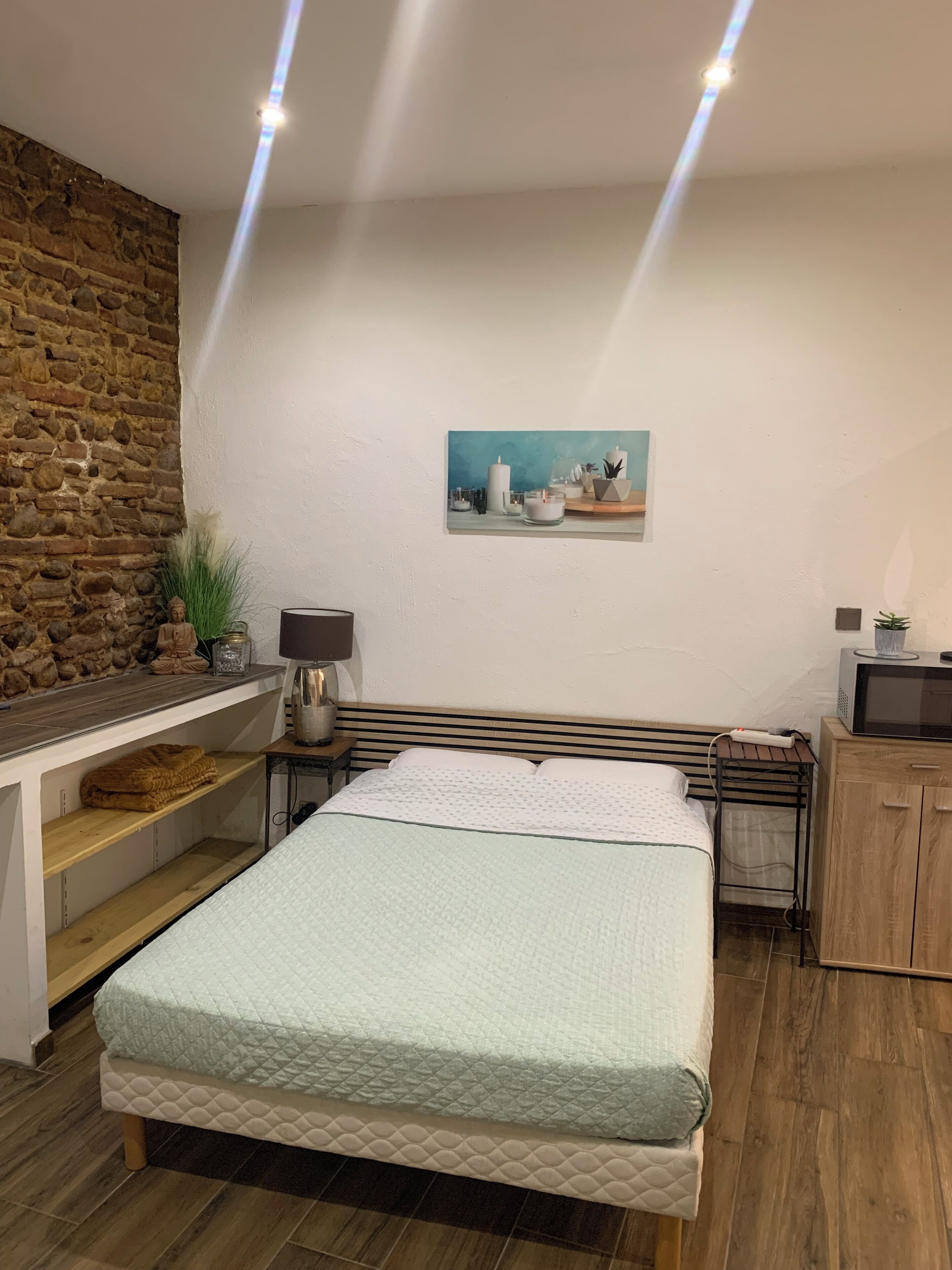 A bedroom with a bed, a desk, a chair, a nightstand, a lamp, a painting, and a stone wall.