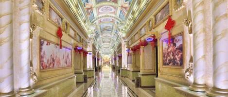 A long, ornate hotel corridor with marble columns, a tiled floor, and a ceiling with painted artwork.