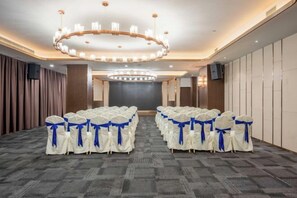 Une salle de conférence avec des rangées de chaises recouvertes d’un tissu blanc et bleu, un grand lustre et un écran de projection à l’avant.
