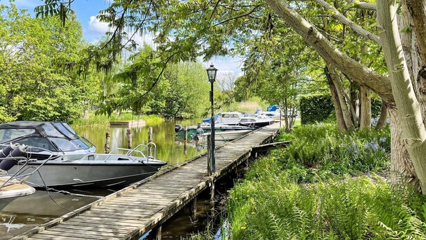 Une jetée en bois s’étendant sur une étendue d’eau calme, bordée de bateaux et entourée d’une végétation luxuriante et d’arbres.