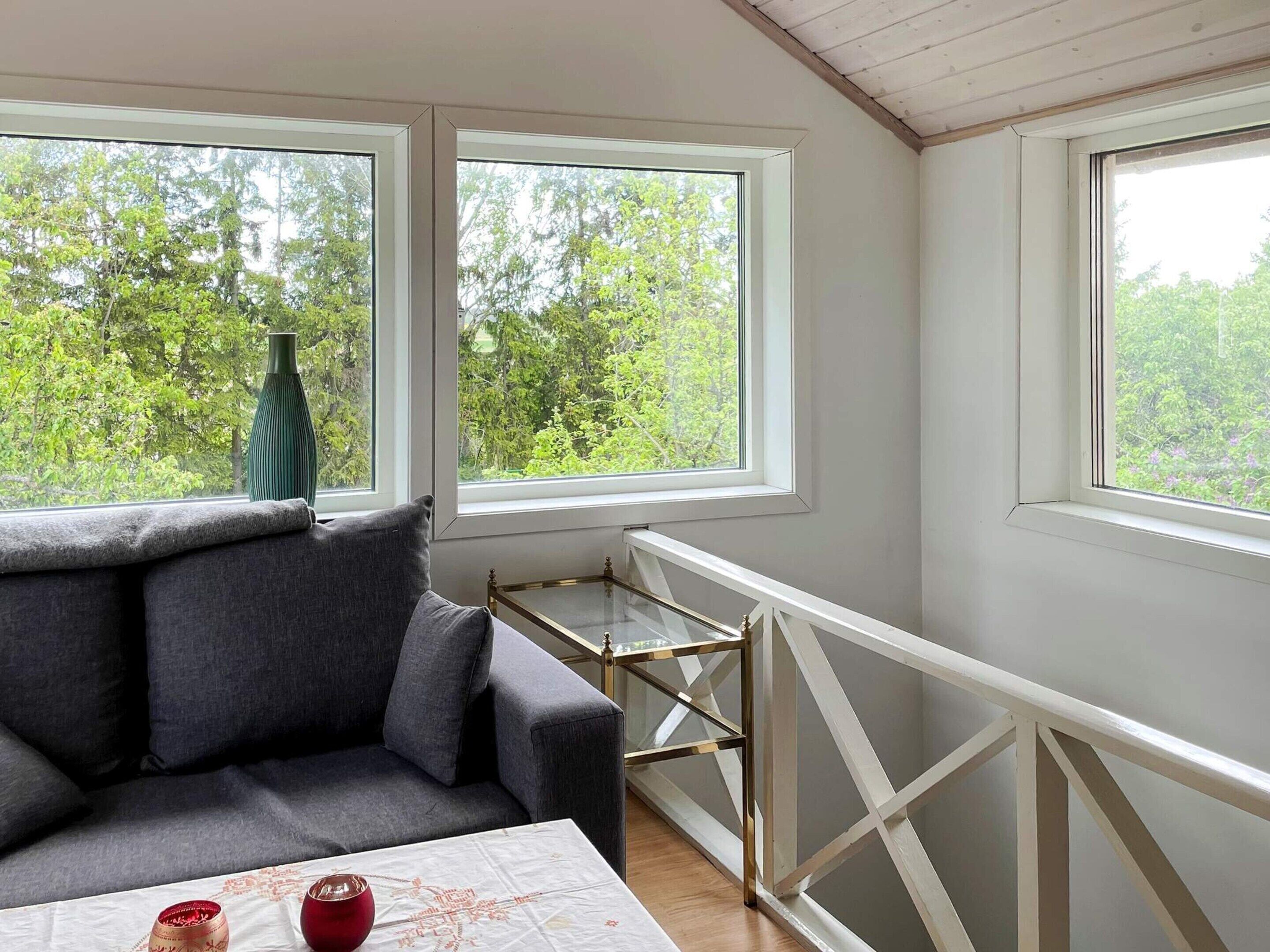 Et værelse med en grå sofa, et lille bord med hvid dug og to vinduer med udsigt til grøn natur.