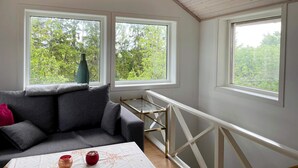 Une pièce chaleureuse avec un plafond en pente, un canapé gris, une petite table et deux fenêtres donnant sur un paysage verdoyant.