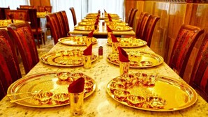 Une longue table à manger, ornée d’assiettes et de verres dorés, entourée de chaises en bois.