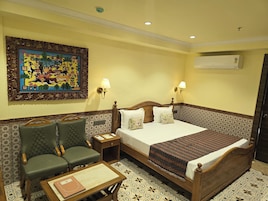 A hotel room with a bed, two chairs, a small table, a nightstand, a wall-mounted lamp, and a decorative wall piece.