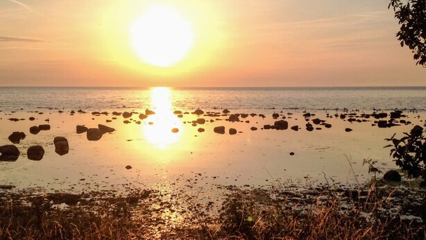Auringonlasku kivikkoisella rannalla, jossa tyyni vesi heijastaa auringonvaloa.