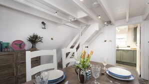 A dining area with a wooden table set for two, white chairs, a vase with flowers, and a view of the kitchen through an open door.