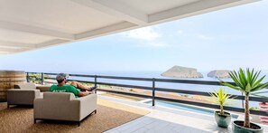 A man relaxing on a sofa with a view of the sea and a balcony with potted plants.