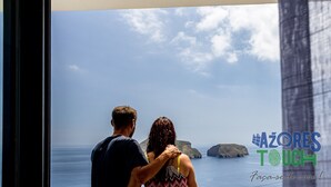 Ein Paar blickt aus einem Fenster auf die Küstenlandschaft. Am unteren rechten Rand ist das Logo 'Azores Touch' zu sehen.