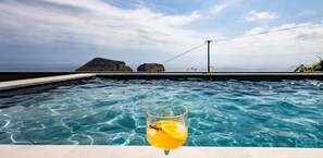A swimming pool with a glass of orange juice in the foreground, a clear sky, and distant rocky formations.
