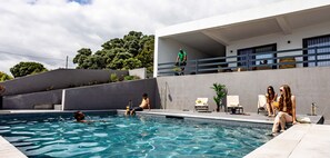 A swimming pool with people relaxing and a person standing on the balcony.