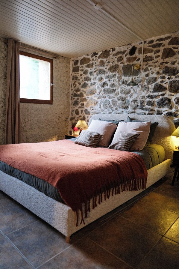 A bedroom with a stone wall, a bed with multiple pillows, a nightstand with a lamp, and a window with curtains.