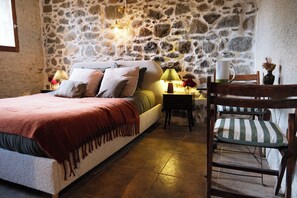 Une chambre avec un mur en pierre, un lit recouvert d’une couverture rouge, une table de chevet avec une lampe et une chaise avec un coussin à rayures.