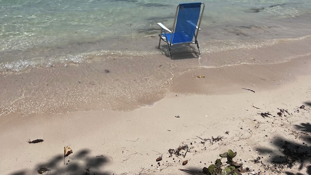Ein Strand mit klarem, seichten Wasser, einem einzelnen blauen Liegestuhl und einer entfernten Küste.