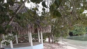 Ein Strandbereich mit einer strohgedeckten Struktur, sandigem Untergrund und umgebender Vegetation.