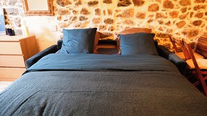 Un lit recouvert d’une couvre-lit bleu foncé et agrémenté d’oreillers, une table de chevet en bois et, en arrière-plan, un mur en pierre.