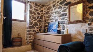 Une pièce aux murs de pierre, avec une commode en bois, une fenêtre avec des rideaux, un miroir et un canapé bleu.