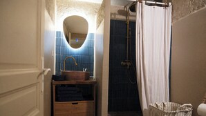 Une salle de bain avec un meuble-lavabo en bois, un miroir, un mur recouvert de carreaux bleus, une douche avec un rideau et une poubelle à rayures.