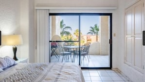 Habitación de hotel con cama, mesita de noche, lámpara, balcón con mesa y sillas, y vista a palmeras.
