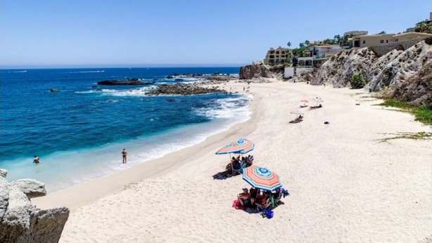 Una playa con agua azul cristalina, algunas sombrillas y un edificio al fondo.