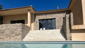 Une maison moderne avec une façade en pierre, une piscine et un ciel d’un bleu éclatant.