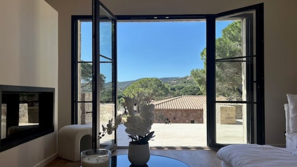 Une chambre avec un lit, une table de chevet sur laquelle est posée une bougie, une chaise et une vue sur une terrasse extérieure, accessible par de grandes portes vitrées.