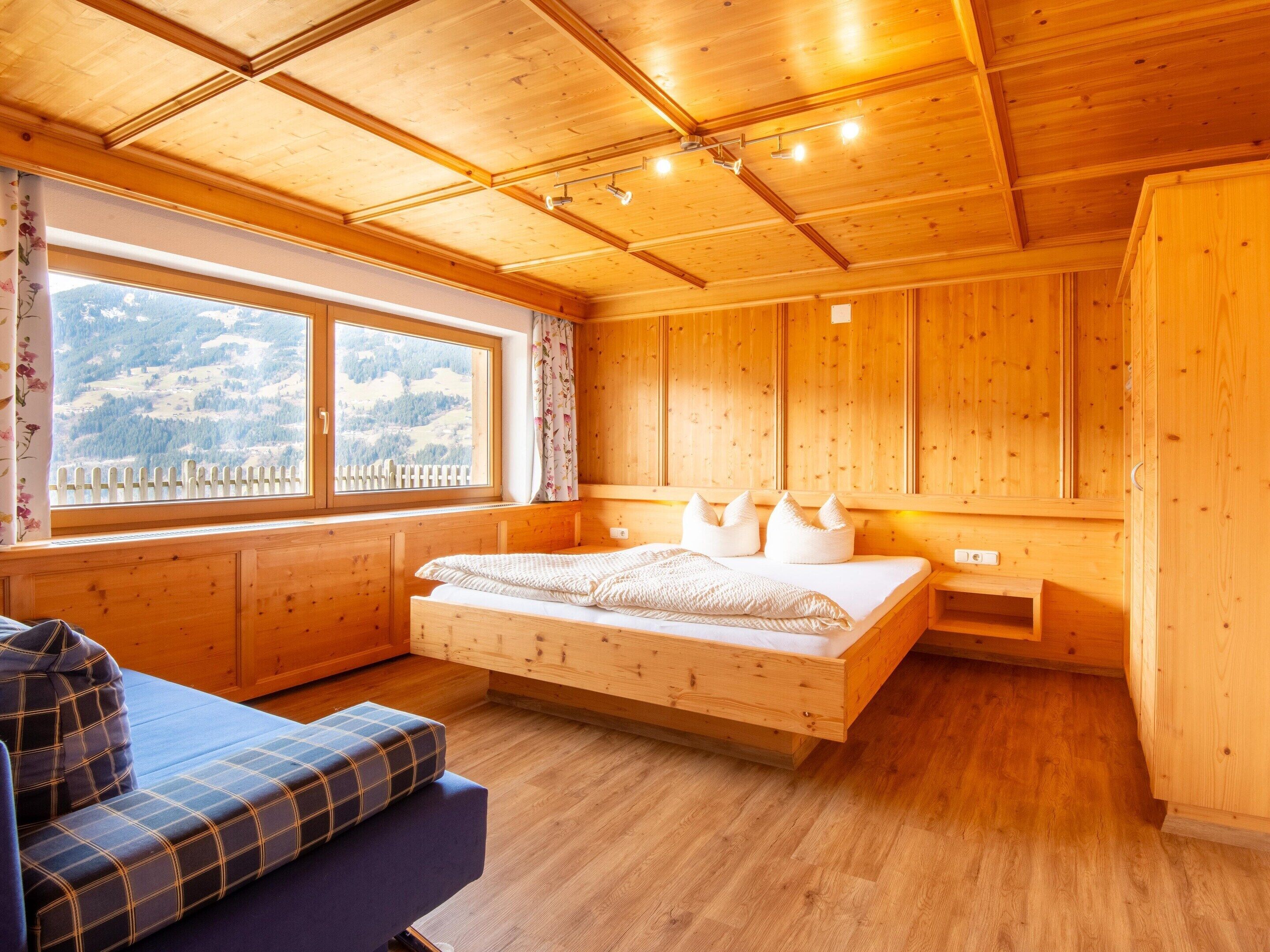 A wooden cabin room with a bed, a window with a view of the mountains, and a sofa.