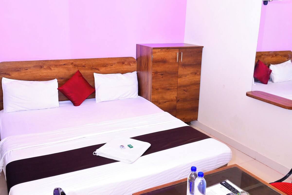 Una habitación de hotel con una cama, un armario de madera, una mesita de vidrio con artículos de aseo y una silla roja.