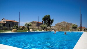 Une piscine à l’eau d’un bleu cristallin, entourée de bâtiments en pierre et d’un arbre.
