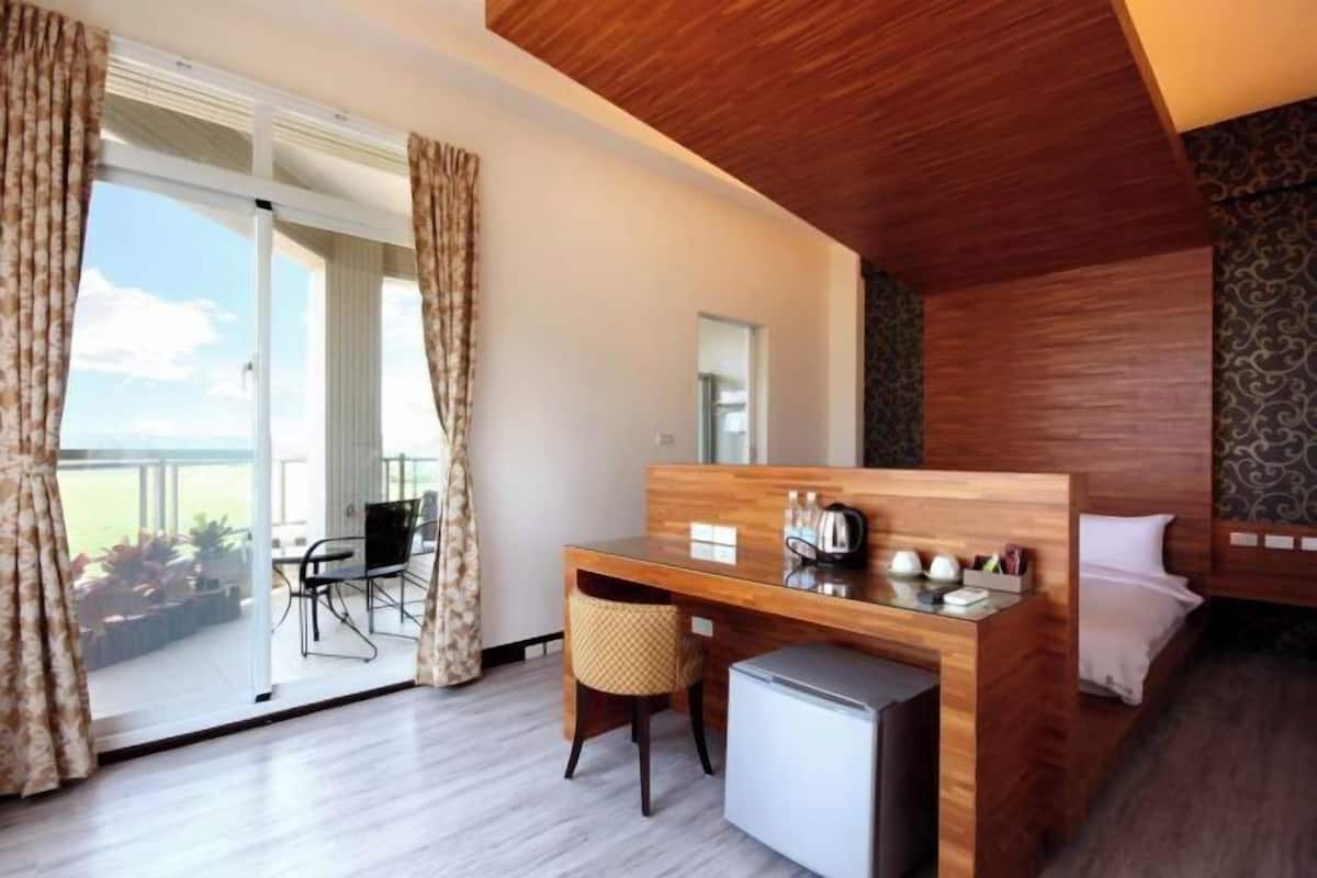 Une chambre d’hôtel avec un bureau en bois, un petit réfrigérateur, un lit avec du linge de maison blanc et un balcon équipé d’une table et de chaises.