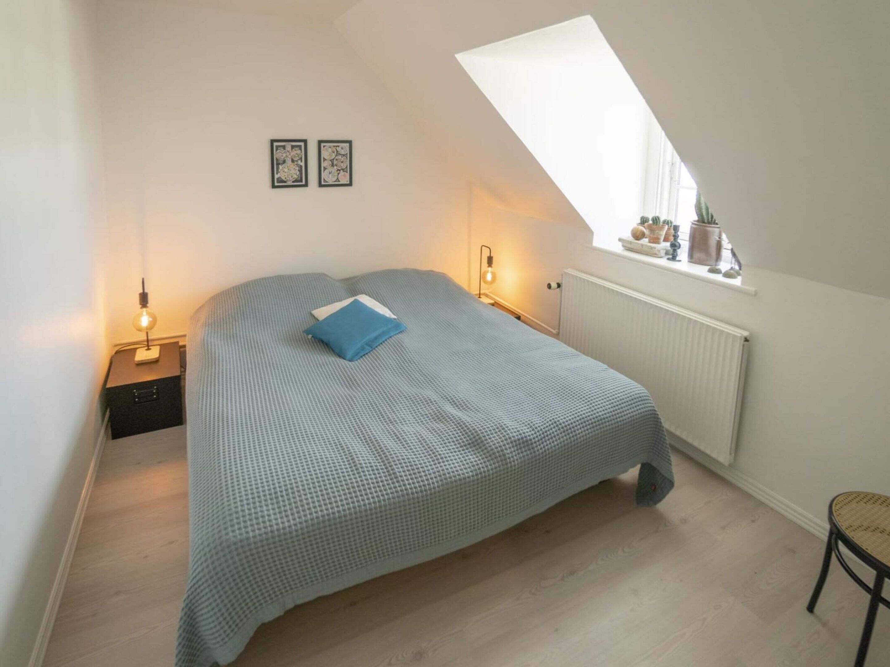 Une chambre avec un plafond en pente, un lit avec un oreiller bleu, deux lampes fixées au mur, une fenêtre donnant sur un paysage agréable et un radiateur.