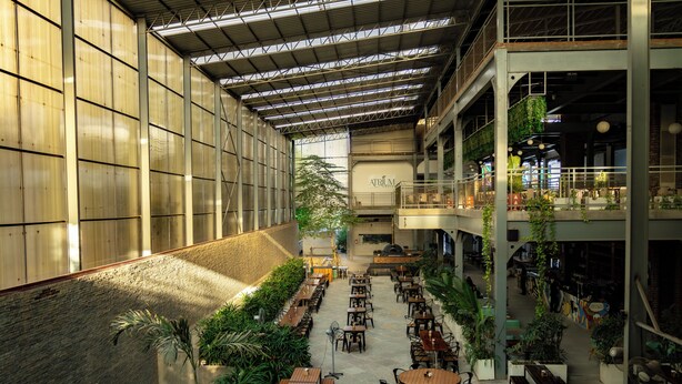 Un vaste espace intérieur doté de nombreuses tables et de bancs, entouré de verdure et surmonté d’un haut plafond agrémenté de lucarnes.