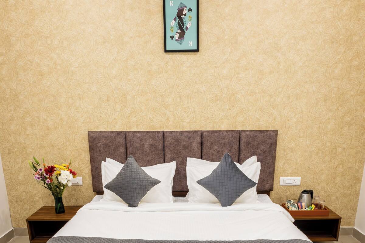 Quarto de hotel com uma cama grande, duas poltronas, uma pequena mesa de madeira com frutas e uma mesa de cabeceira com um vaso de flores.